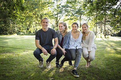 small group of teenagers sitting together on a slackline in a park